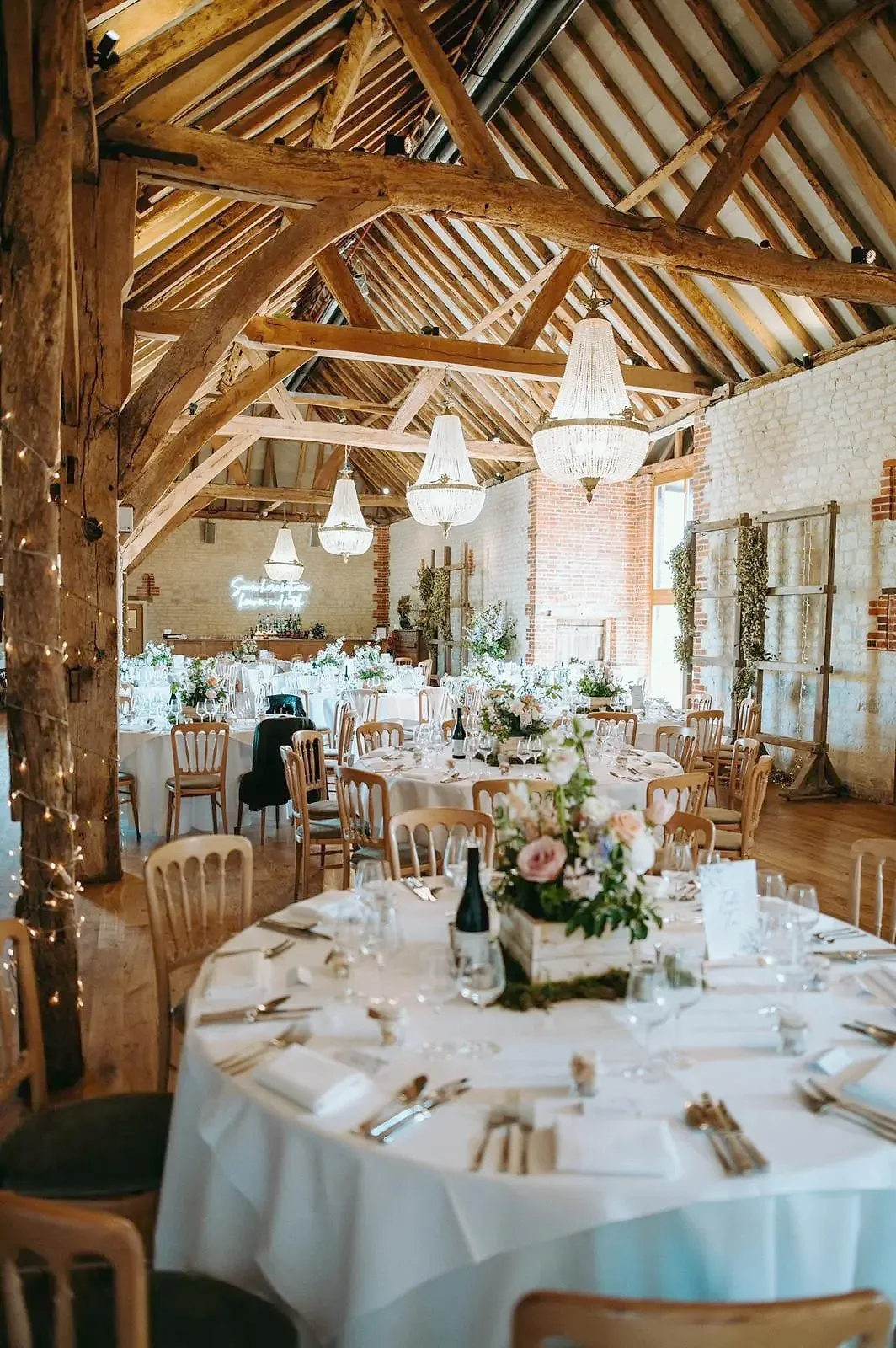 The barn laid for dinner with round tables, white linen, flowers and crystal chandeliers under the oak trusses