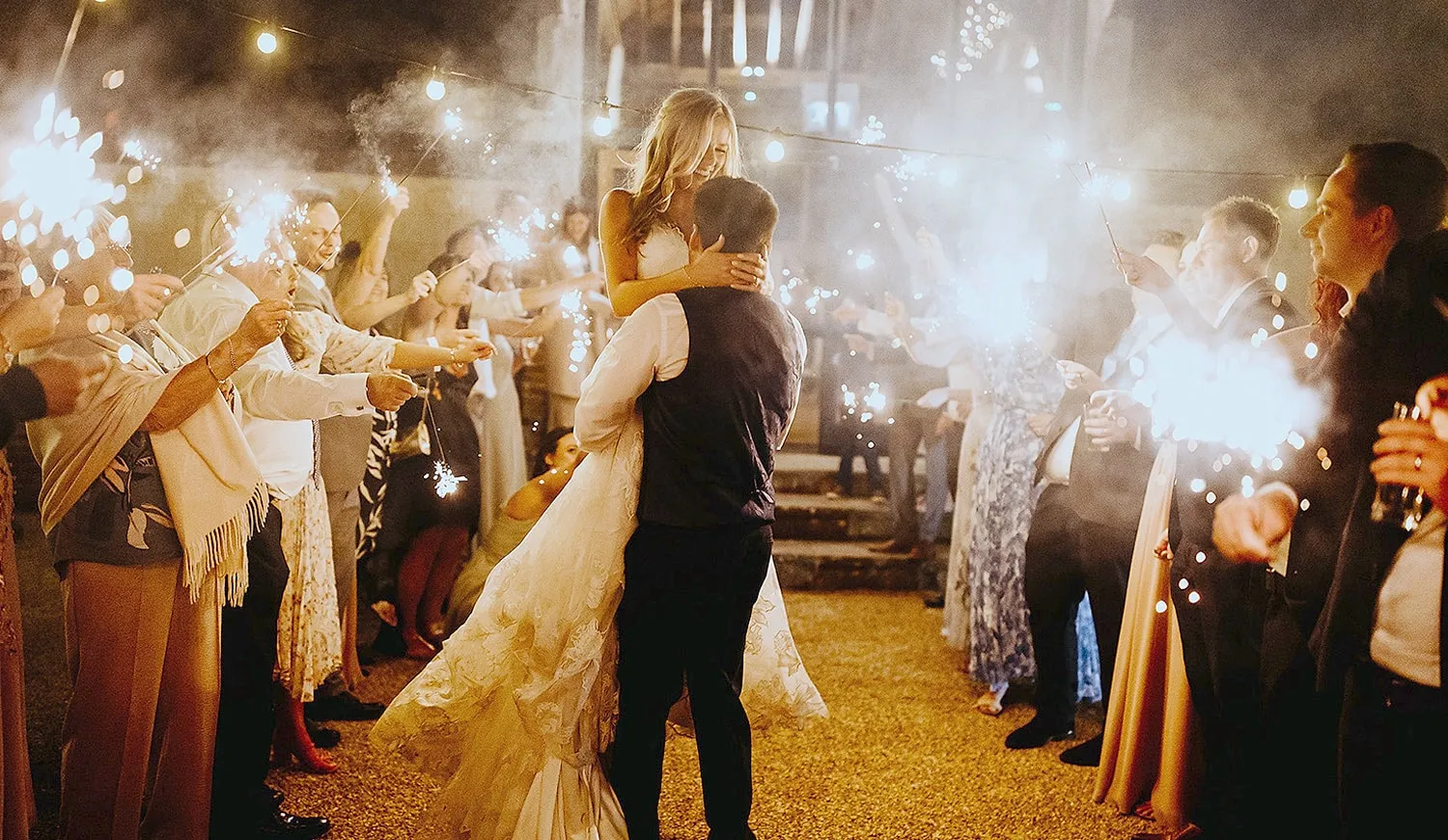 Guests holding sparklers as the groom lifts the bride in the courtyard at night